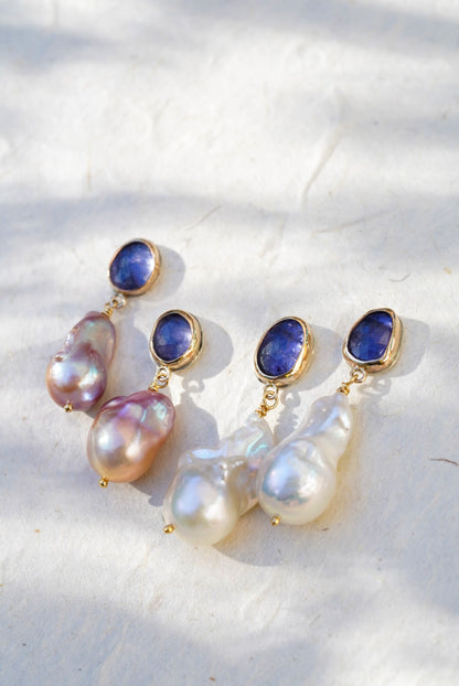 Four gold drop earrings with oval tanzanite gemstones at the top and irregular baroque pearl drops below, displayed  against a light textured background.