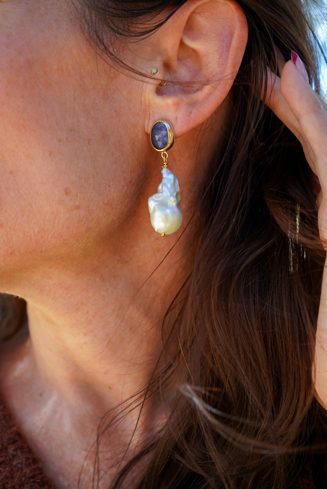 A close-up of a woman's ear wearing a gold dangle earring with a tanzanite oval stone and a large baroque pearl drop.