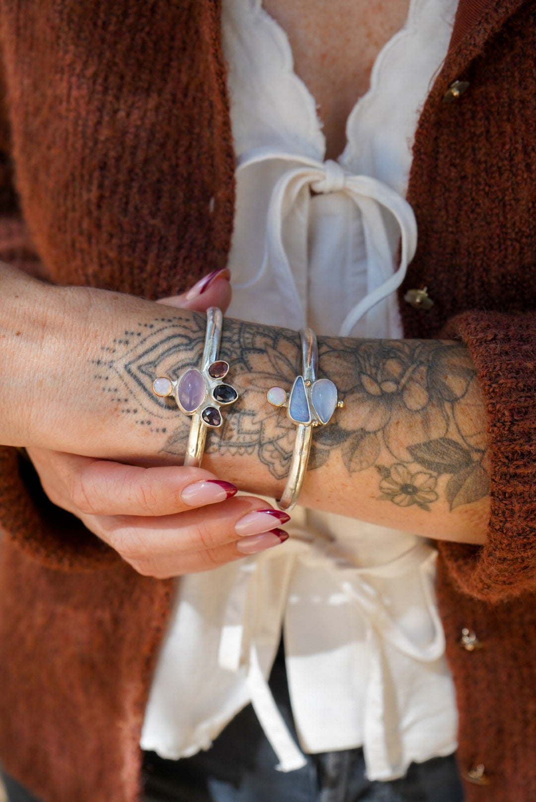 A close-up of a person's tattooed wrist wearing two silver cuff bracelets with organic-shaped pastel gemstones, styled with a white blouse and brown cardigan.