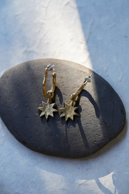 A pair of bronze hoop earrings with dangling star charms, displayed on a dark smooth stone against a light textured background.