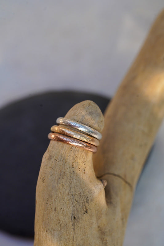 Three thin textured metal rings in silver, gold, and rose gold tones, displayed on a piece of driftwood.