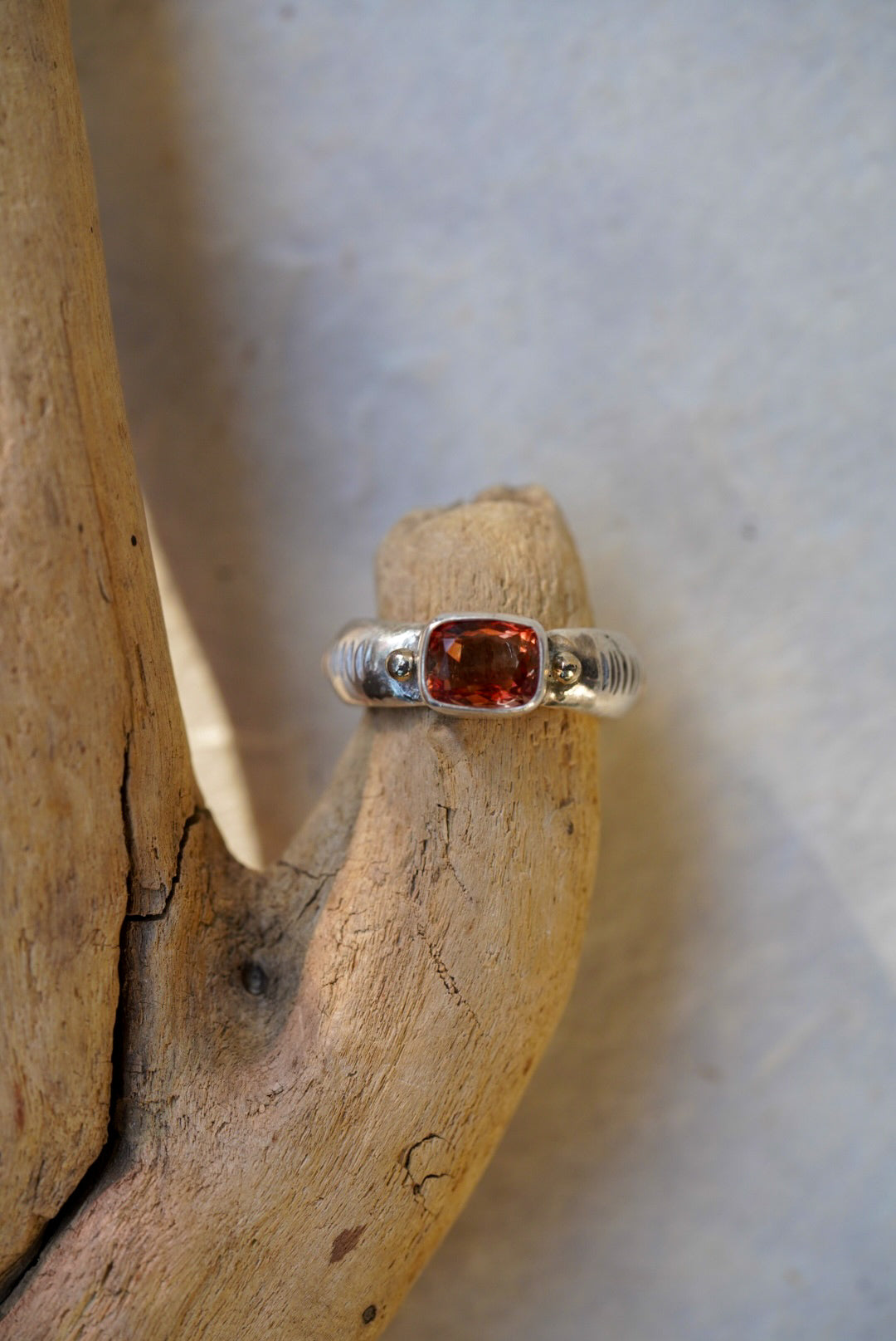 A silver ring with a cushion-cut orange-red gemstone, displayed on a piece of natural driftwood against a light gray background.