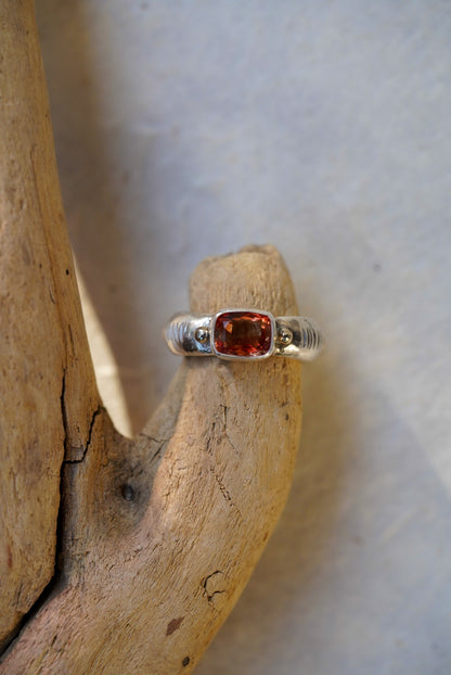 A silver ring with a cushion-cut orange-red gemstone, displayed on a piece of natural driftwood against a light gray background.