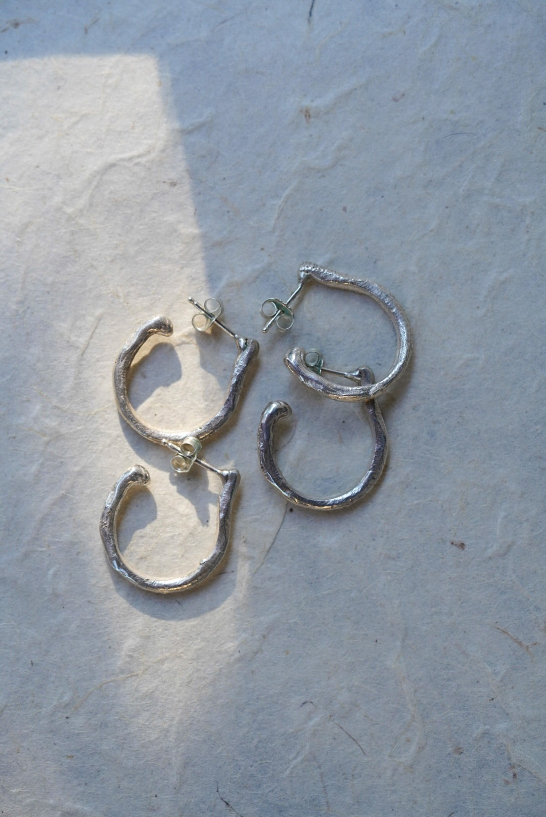 A pair of textured silver hoop earrings with post-back closures, displayed on a light gray textured paper background.