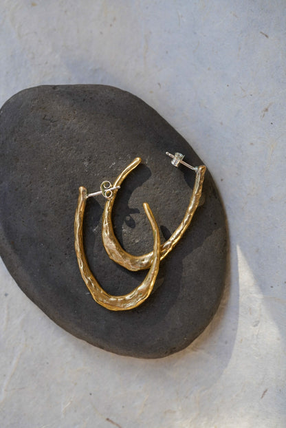 A pair of organic textured gold crescent hoop earrings with silver posts, displayed on a dark gray stone against a light gray background.