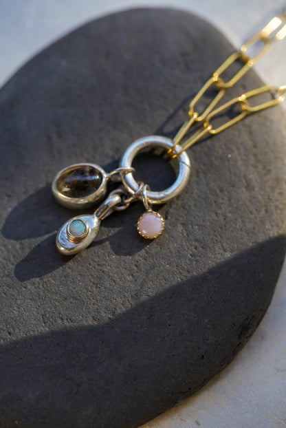A close-up of a gold and silver pendant necklace resting on a dark stone. The necklace features a silver ring with dangling charms, including a silver teardrop with an opal stone, a silver oval with a rutilated quartz, and a small pinkish-orange opal charm.