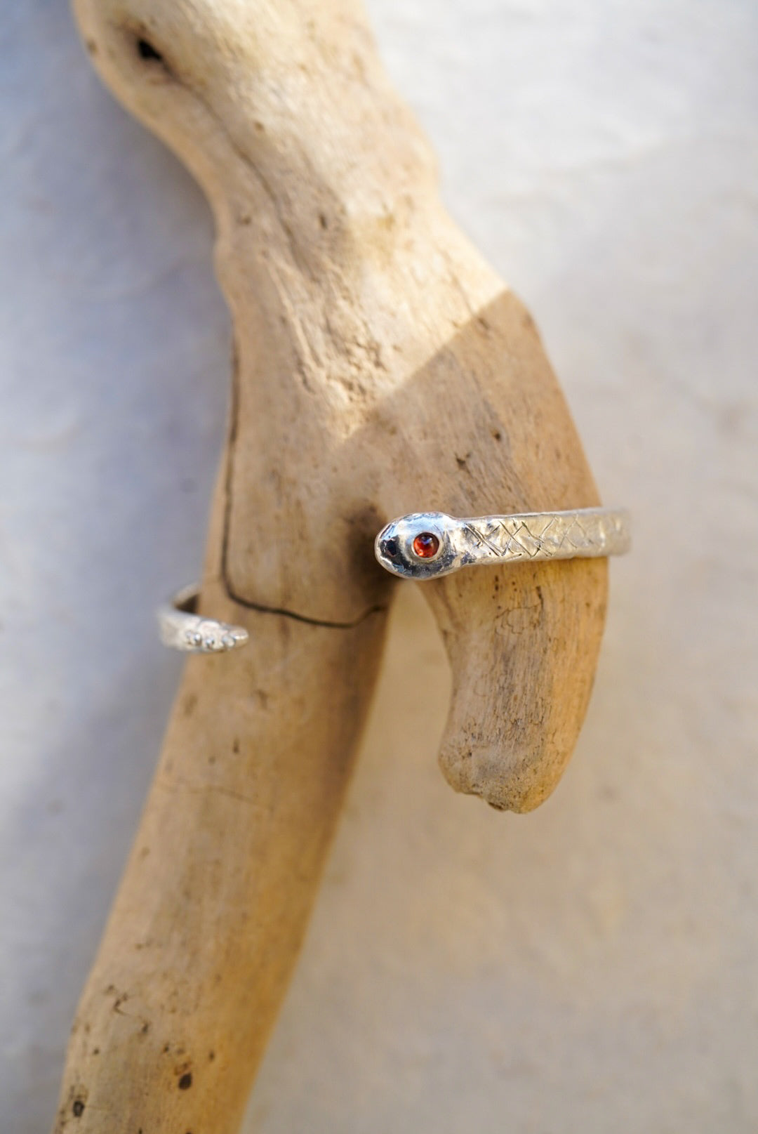 A silver snake cuff with a small garnet, resting on a piece of driftwood against a light gray background.