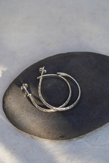 A pair of textured silver hoop earrings resting on a smooth, dark gray stone against a light beige background.