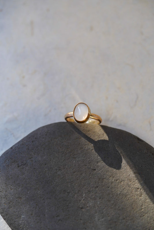 A gold ring with an oval milky-purple cabochon stone, displayed on a dark gray stone prop against a light beige textured background.