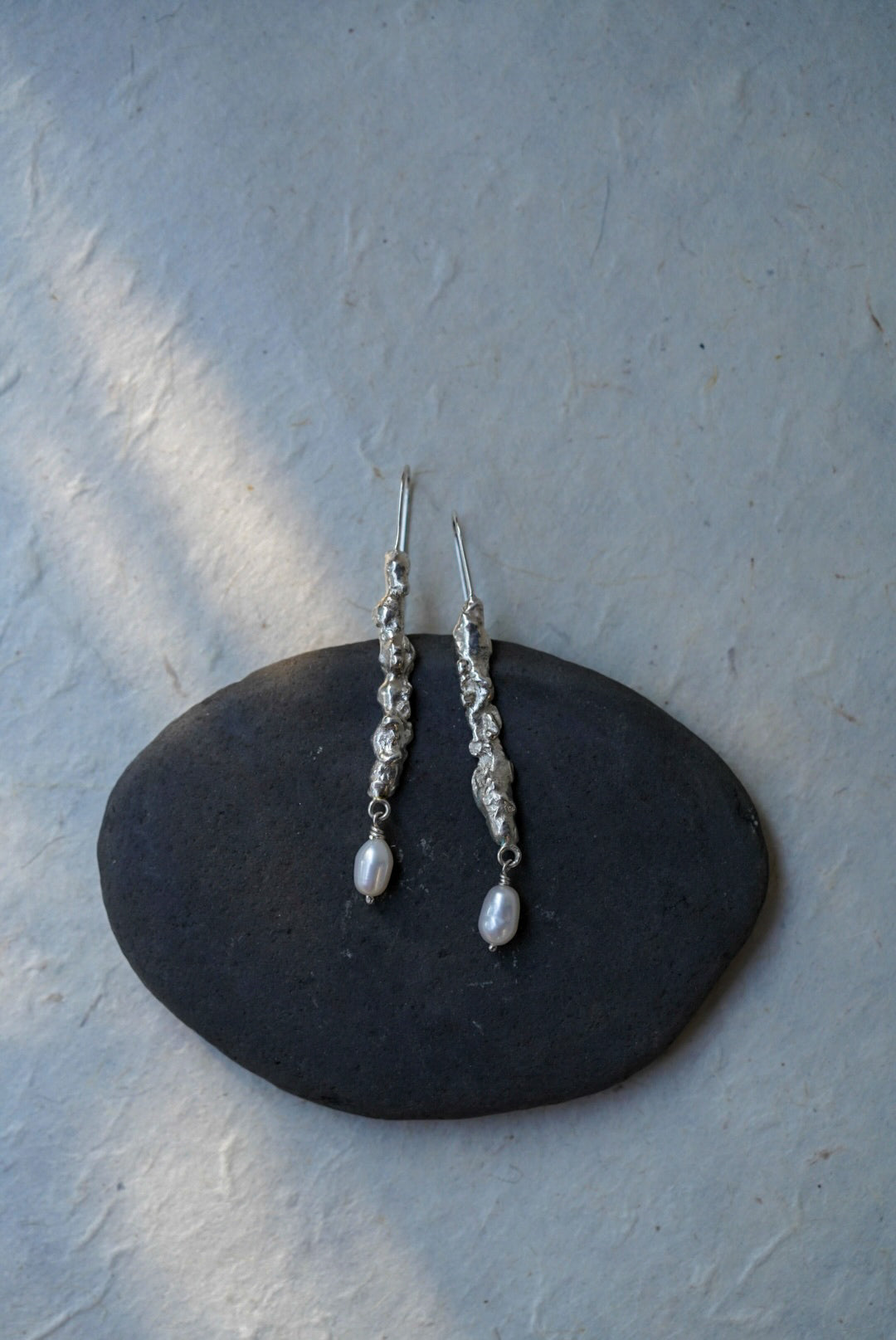 A pair of textured recycled silver dangle earrings and small white pearl-like drops, displayed on a dark oval stone against a light textured wall.