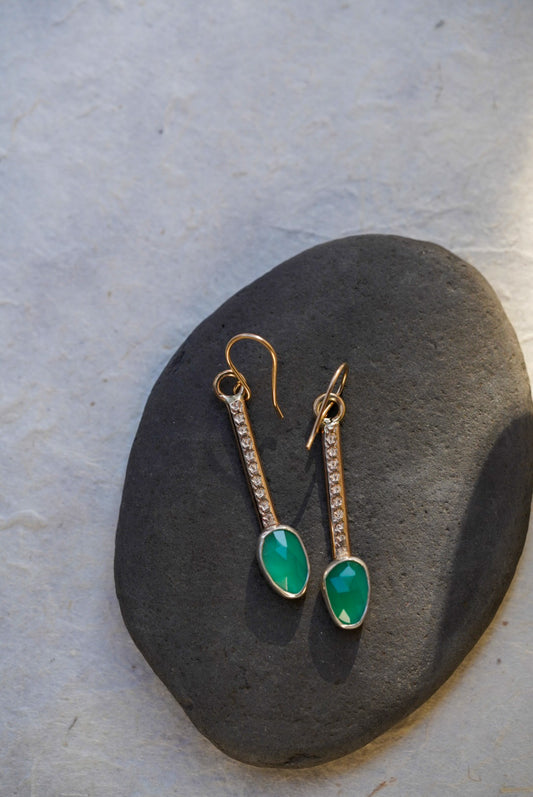 A pair of gold drop earrings with marquise-cut emerald green chalcedony, displayed on a dark gray stone against a light background.