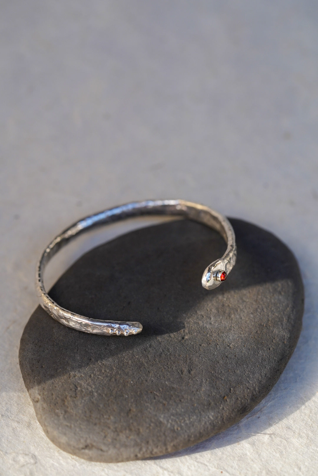 A silver snake cuff with a small garnet, resting on a dark stone against a light gray background.