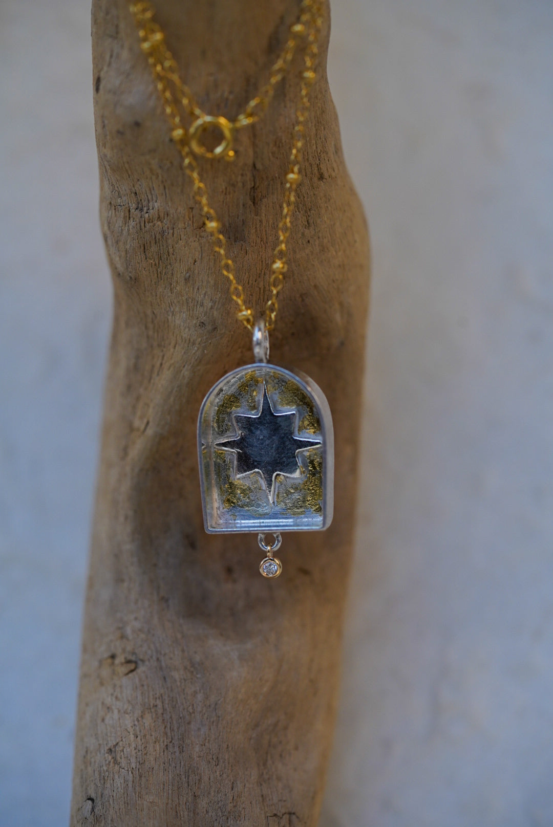 A gold chain necklace with a arched pendant containing a silver star with a dusting of gold, displayed on a piece of driftwood.