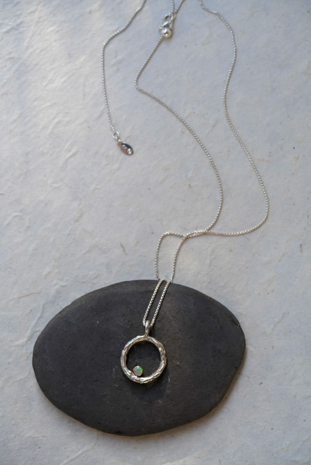 A silver chain necklace with a textured circular pendant resting on a dark stone, set against a light textured background.