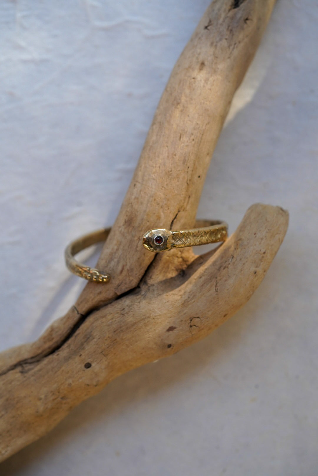A bronze snake cuff with a small garnet, wrapped around a piece of natural driftwood on a light textured background.