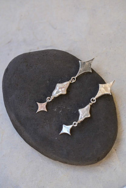 A pair of silver dangle earrings with star-shaped and star pearls, displayed on a dark stone against a light beige background.