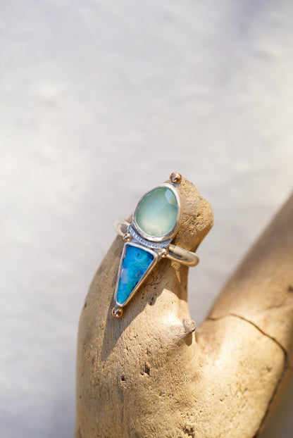 A silver ring with a pale blue stone and a triangular bright blue opal stone, displayed on a textured tan stone stand.