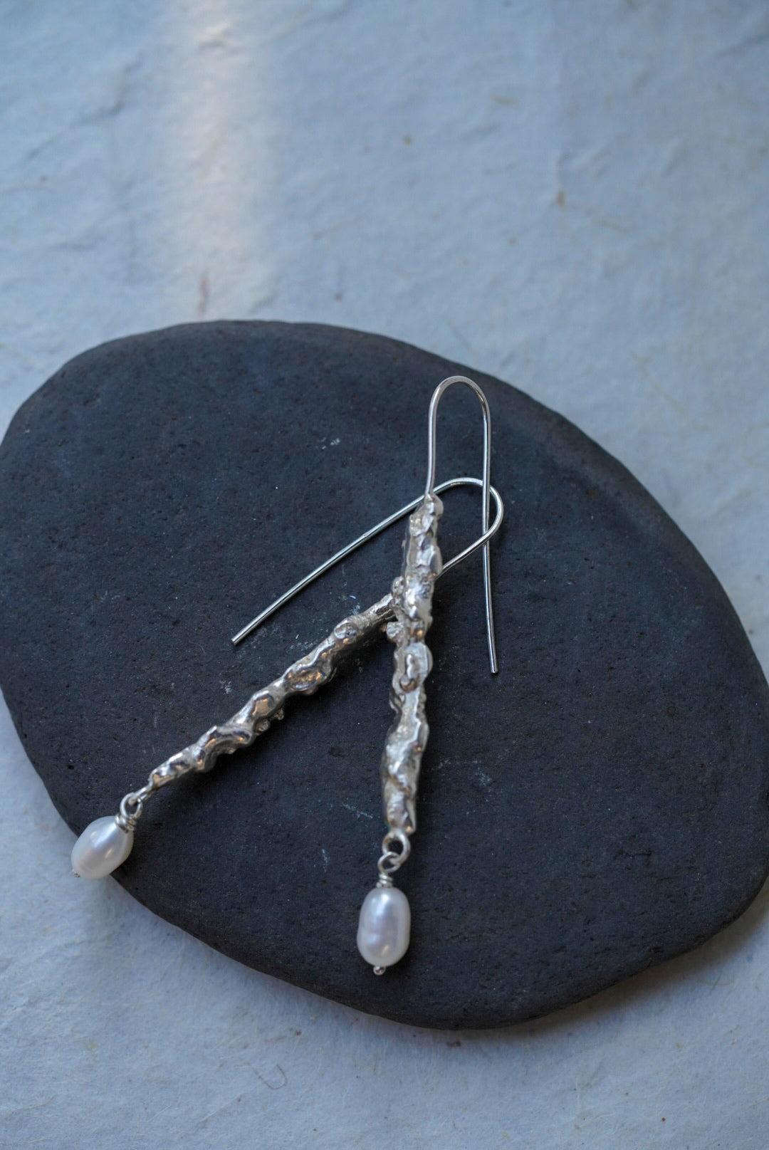 A pair of handmade textured recycled silver dangle earrings and small white pearl-like drops, displayed on a dark oval stone against a light textured wall.