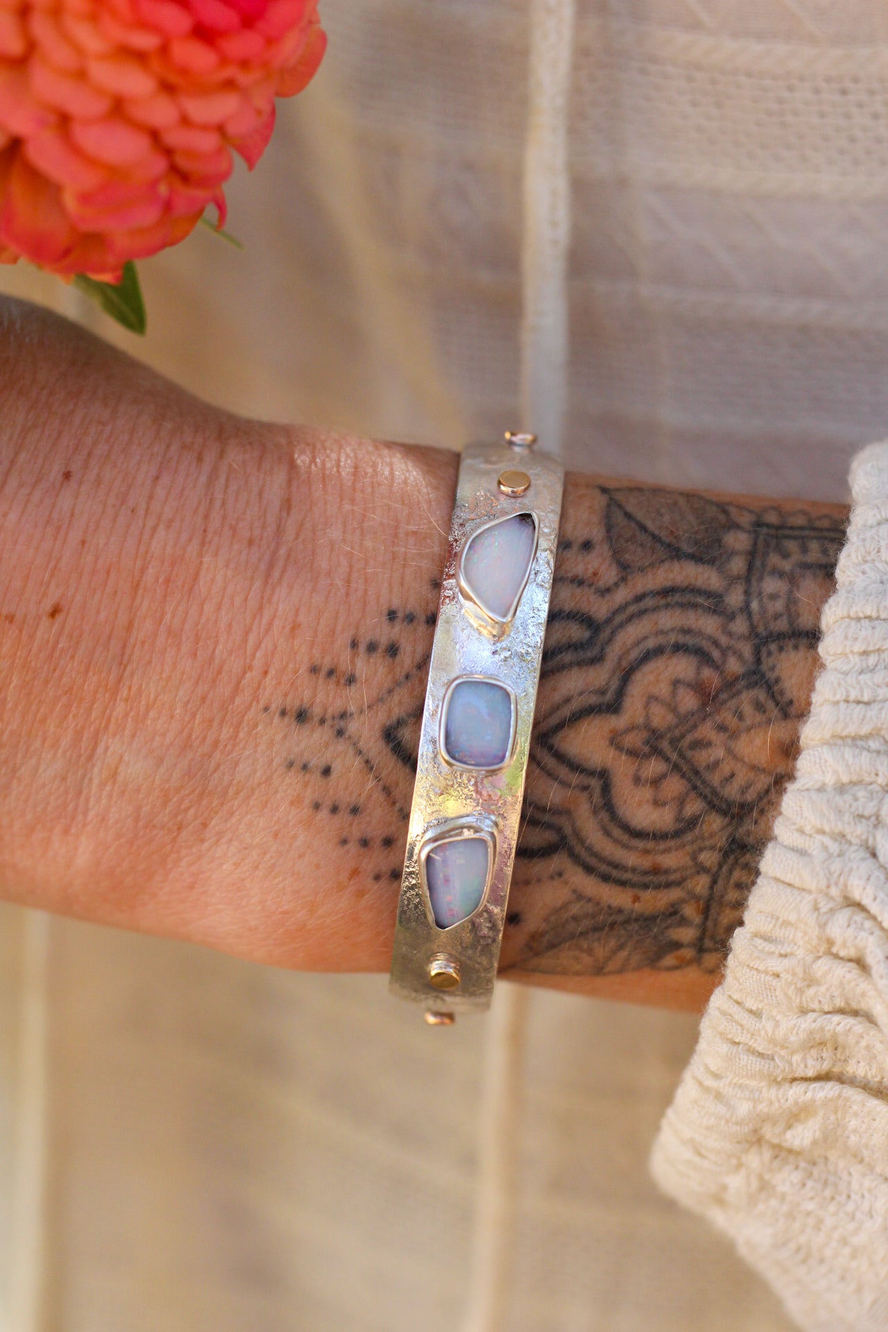 Silver bracelet with three Australian opal stones on a wrist with tattoos, next to a orange flower.
