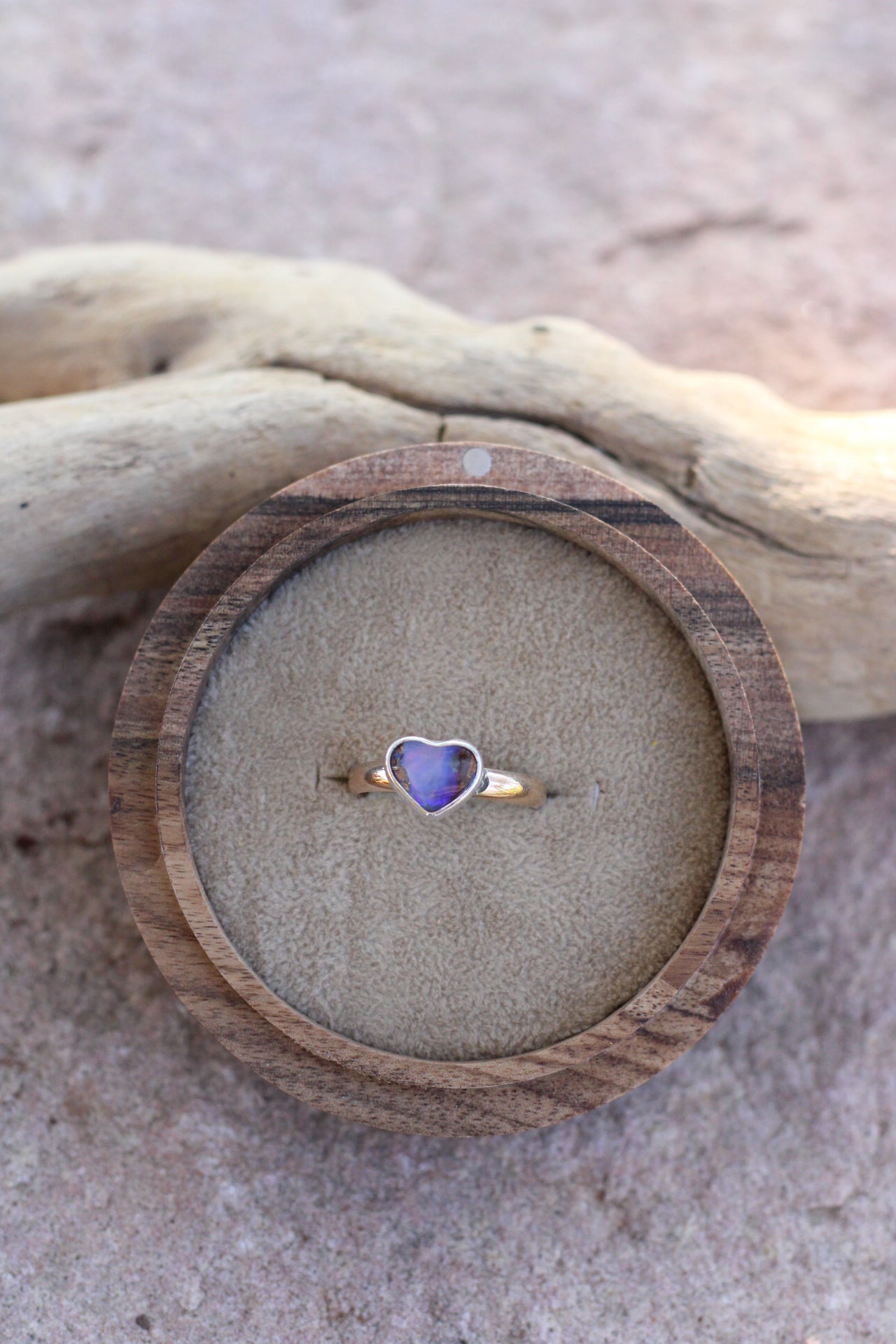 Heart-shaped opal ring with a purple stone in a wooden box on a sandy surface
