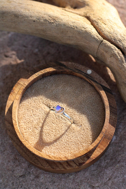 Wooden ring box with a heart shaped  opal ring inside, placed on sand with driftwood.