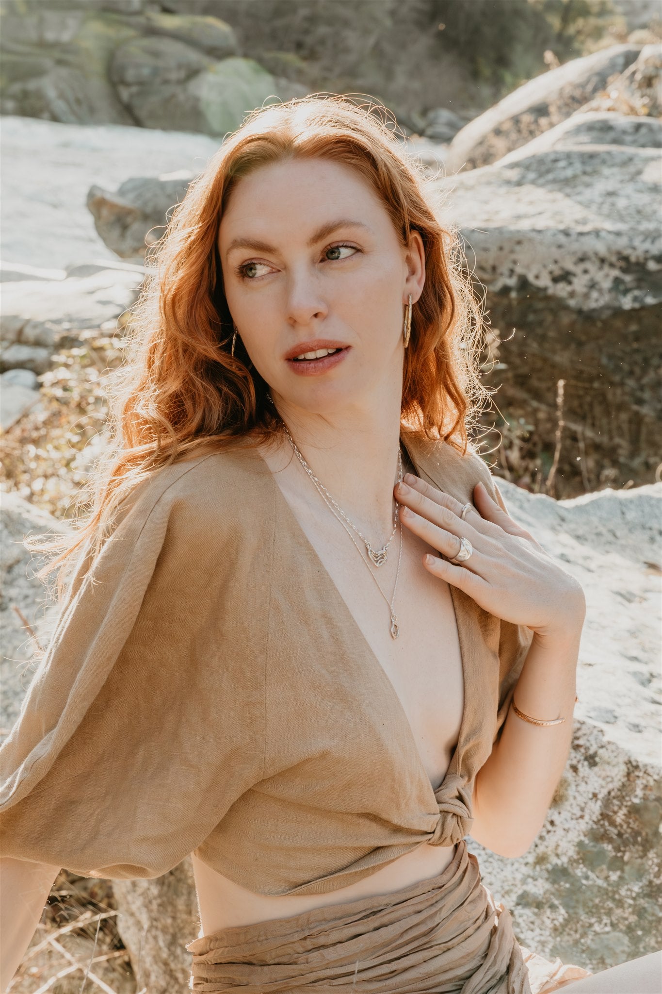 Woman with long hair standing outdoors near rocks, wearing jewelry
