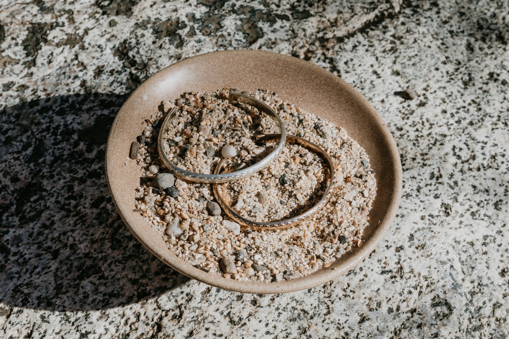 Two bracelets on a small ceramic plate with sand, placed on a textured stone surface.