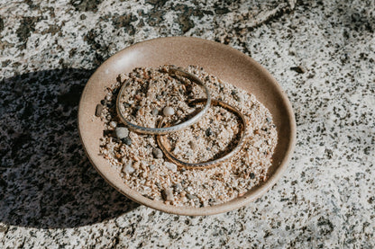Two bracelets on a small ceramic plate with sand, placed on a textured stone surface.