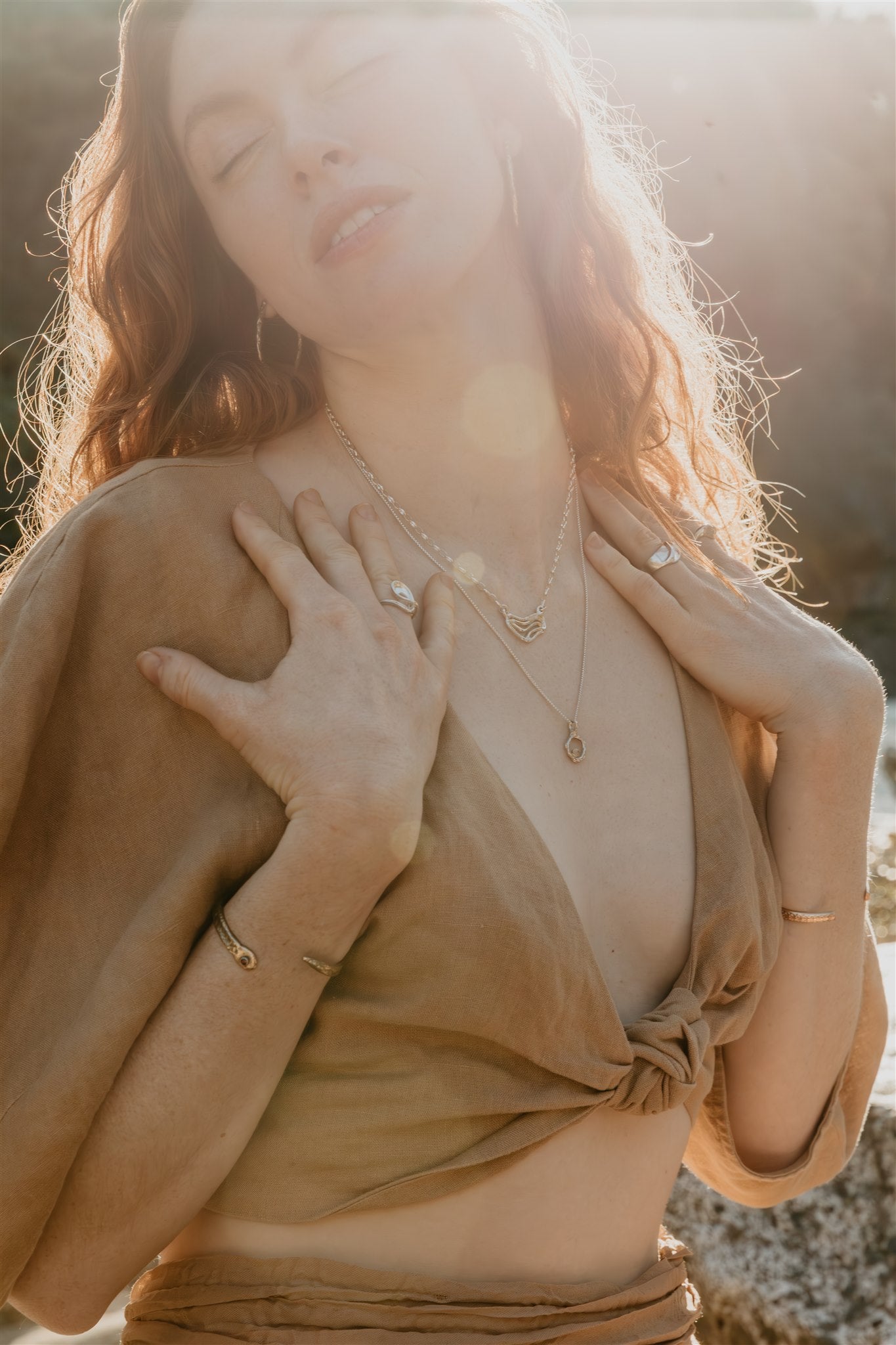 Woman with closed eyes wearing a beige top and gold and silver jewelry against a blurred natural background