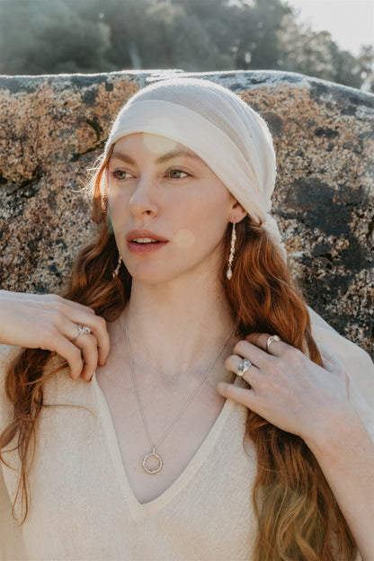 Woman with long hair wearing a white headscarf and beige sweater against a stone wall.