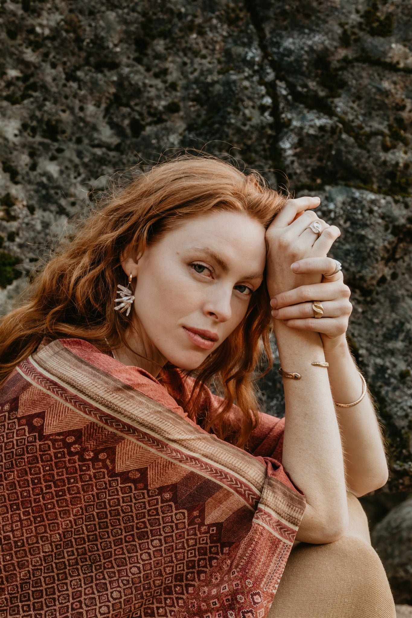 Woman with patterned shawl with earrings, rings, and bracelets against a stone 