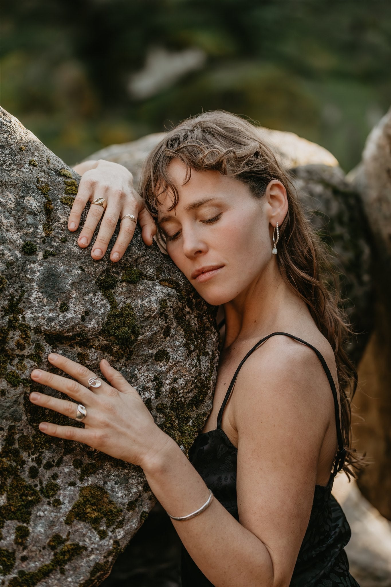 Woman in a black dress leaning against a rock with a blurred natural background, wearing jewelry