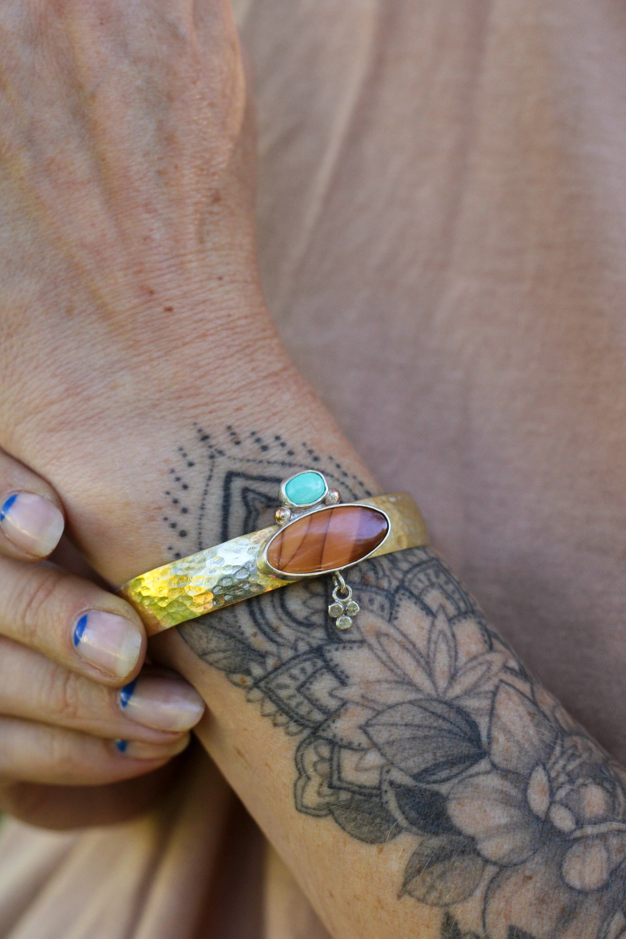 Close-up of a hand wearing a gold bracelet with a brown and turquoise stones on a tattooed arm.