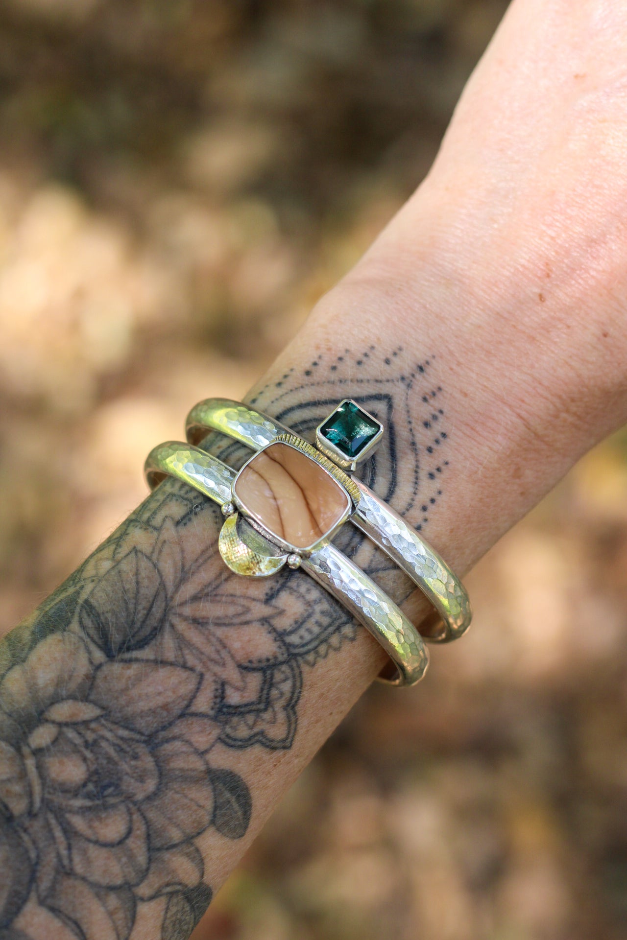 Close-up of a wrist wearing a statement cuff with a brown and green gemstone against a blurred natural background.