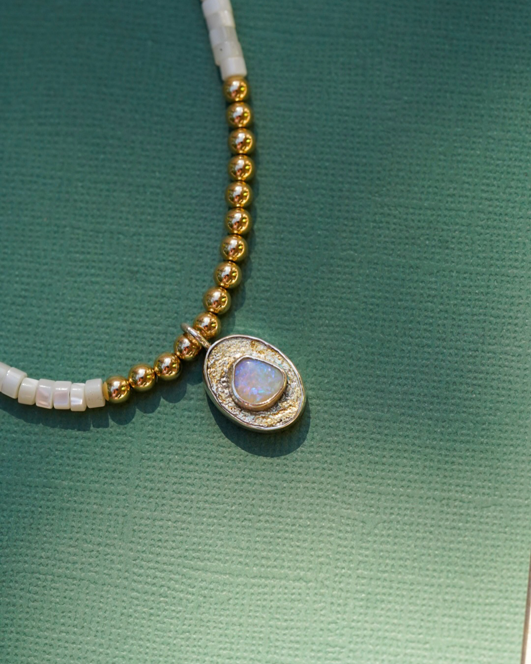 A close-up of a beaded necklace featuring pearl beads, gold round beads, and a central oval pendant with a opal iridescent stone, set against a textured green background.