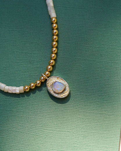 A close-up of a beaded necklace featuring pearl beads, gold round beads, and a central oval pendant with a opal iridescent stone, set against a textured green background.